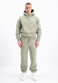 Set van olijfgroene hoodie en joggingbroek, gemaakt van zacht materiaal. Met een voorvak en logodetail. Gecombineerd met witte sneakers.