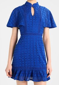 Vestido de encaje azul con un alto cuello, detalle de ojal, mangas cortas acampanadas y un dobladillo con volantes. Presenta patrones intrincados y una tela texturizada.