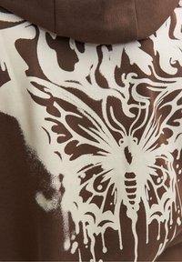 Felpa marrone con una grande grafica di una farfalla bianca caratterizzata da dettagli intricati, posizionata su uno sfondo in tessuto texture.