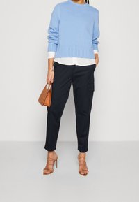 Lichtblauwe gebreide trui met geribbelde zoom, laag over een wit shirt. Donkerblauwe cargobroek en tan hakken maken de outfit compleet.