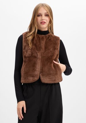 Donna che indossa un gilet marrone senza maniche in ecopelliccia sopra una maglia nera a maniche lunghe e pantaloni neri, in piedi con una mano nella tasca del gilet.