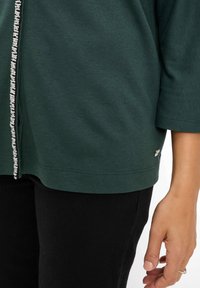 Donkergroene shirt met korte mouwen, voorzien van een knopenlijst en zilveren pailletten langs de voorkant. Zwarte broeken en een zilveren detail op het kledingstuk.