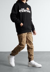 Hoodie noir avec logo "ellesse", pantalon cargo kaki avec plusieurs poches et baskets blanches. La tenue présente une coupe décontractée et des poignets en tricot.