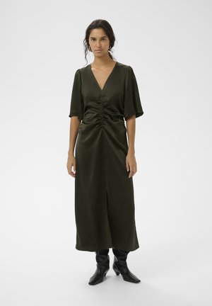 Dunkelgrünes, satiniertes Kleid mit kurzen Puffärmeln, tiefem V-Ausschnitt, Raffung an der Taille und einem Schlitz vorne. Kombiniert mit schwarzen Absatzstiefeln.