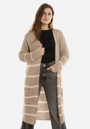 Jonge vrouw in een lange beige gestreepte gebreide cardigan, zwarte top en hoogwaardige vervaagde zwarte jeans, staande met één hand in haar zak.