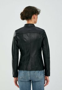 Giacca in pelle nera con vestibilità su misura, dotata di colletto e cuciture verticali. La parte posteriore presenta una leggera arricciatura in vita, abbinata a denim chiaro.