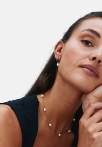 Collar de oro con perlas blancas irregulares, llevado con una blusa oscura. La modelo apoya la mano en el mentón, mostrando el delicado diseño de la joya.