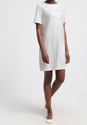 Mujer con un vestido gris claro, texturizado, de manga corta y a la altura de la rodilla, y zapatos planos blancos, de pie contra un fondo blanco liso.