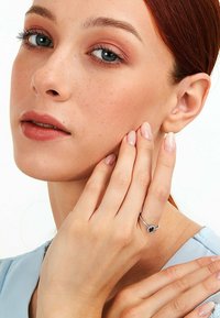 Silberring mit einem dunklen Edelstein, getragen an einer Hand mit natürlichen, polierten Nägeln. Das Modell hat helle Haut und rote Haare. Sanfte, neutrale Kleidung.