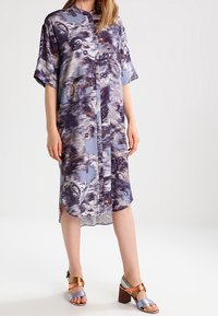 Knopfle dress mit einem blauen und lila abstrakten Muster, kurzen Ärmeln und einem abgerundeten Saum. Kombiniert mit mehrfarbigen Absatzsandalen.