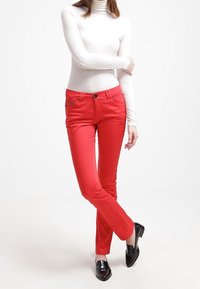 Pantalones ajustados rojos de algodón elástico, con bolsillos laterales y un acabado brillante. Llevados con una blusa blanca de cuello alto y mocasines negros.