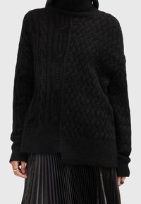 Pull noir en maille avec un col montant, présentant des motifs torsadés et en losange texturés, associé à une jupe plissée noire.