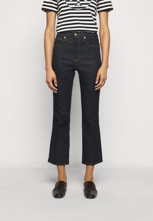 Osoba ubrana w czarne spodnie dzwony z krótką nogawką, czarne mokasyny i czarno-biały pasiasty krótki rękaw, stojąca na jednolitym tle.