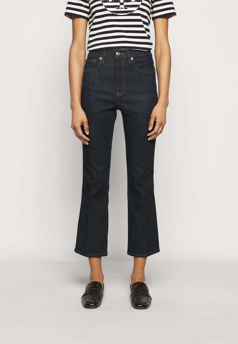 Personne portant un jean noir évasé raccourci, des mocassins noirs, et un haut à manches courtes rayé noir et blanc, debout devant un fond uni.