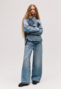 Jeans jakna z lažjim pranjem in velikimi žepi, v kombinaciji z širokimi hlačami. Obe imata raztrgane robove in sproščen kroj. Črne čevlje dopolnjujejo videz.