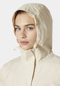Beige waterdichte jas met een hoge kraag en verstelbare capuchon. Voorzien van een ritssluiting aan de voorkant en branding op de borst. Gladde stoftextuur.