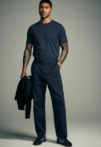 Marineblauwe T-shirt met korte mouwen, gecombineerd met marineblauwe broek. Het model houdt een donker jasje vast en draagt donkere sneakers met groene strepen.