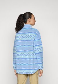 Polarowy pulower w jasnoniebieskim kolorze z geometrycznymi wzorami w ciemniejszym niebieskim, zielonym i różowym. Posiada wysoki kołnierz i luźny krój.