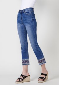 Blå denimjeans med en figurnära design och fransade fållar, med ett färgglatt paisley-mönster vid fållarna och standard fem fickor.