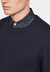 Mężczyzna w ciemnoniebieskim swetrze o ribbowanym splocie, noszący go na niebieskiej koszuli dżinsowej z zapięciem pod szyją.