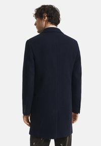 Uomo con capelli ricci che indossa un lungo cappotto blu scuro, di spalle, su uno sfondo bianco semplice.