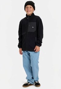 Mustast fliisist jope, millel on tõmblukk ja rinnatasku. Kandes seda helesiniste teksapükste peal ning kombineerituna mustade tenniste ja mütsiga.