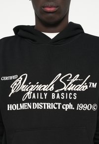Μαύρο hoodie από βαμβακερό μείγμα, με λευκό κείμενο: "CERTIFIED Originals Studio™ DAILY BASICS HOLMEN DISTRICT cph. 1990©".
