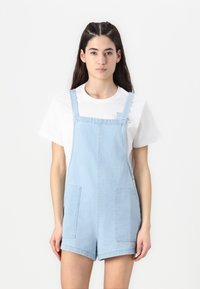 Világoskék denim overál állítható pántokkal, két elülső zsebbel és laza fit-zel. Alatta egy fehér pólóval párosítva.