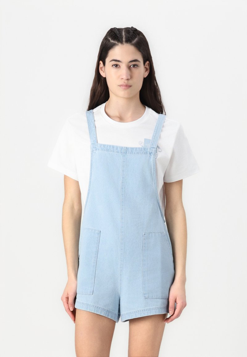 Világoskék denim overál állítható pántokkal, két elülső zsebbel és laza fit-zel. Alatta egy fehér pólóval párosítva.