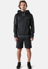 Svart atletisk hoodie med känguruficka, dragsko vid huvan och logotyp. Matchad med svarta knälånga shorts, bärs med svarta sneakers.