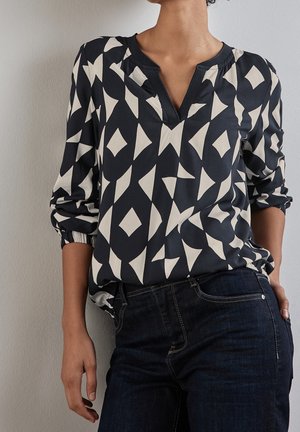 Vrouw draagt een zwarte blouse met witte geometrische patronen en donkerblauwe spijkerbroek, staand tegen een effen lichte muur.