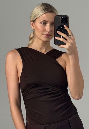 Blondynka z niskim kucykiem, ubrana w czarny, marszczony top bez rękawów, robi sobie selfie w lustrze czarnym telefonem na szarym tle.