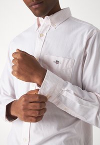 Camicia button-up a righe rosa chiaro con colletto, tasca frontale e logo ricamato. Il tessuto appare liscio con una vestibilità classica.