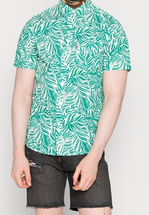 Homme portant une chemise à manches courtes boutonnée à motif de feuilles tropicales vertes et blanches et un short en denim noir usé.