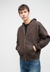 Καφέ φούτερ με κλείσιμο μπροστά και κουκούλα με κορδόνι, καγκουρό τσέπες και ένα μικρό λογότυπο. Φορεμένο πάνω από μαύρο μπλουζάκι και συνδυασμένο με μπλε τζιν.