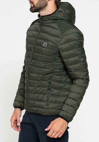 Giacca imbottita verde scuro con cappuccio, dotata di zip, tasche laterali e un logo minimalista sul petto. Tessuto liscio e design leggero.