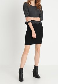 Robe noire avec une silhouette ajustée, associée à un top court noir et blanc à pois. Mannequin portant des bottines noires à la cheville. Textures lisses.