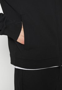 Main partiellement dans la poche d'une veste noire zippée portée sur une chemise blanche et un pantalon noir, sur un fond uni.