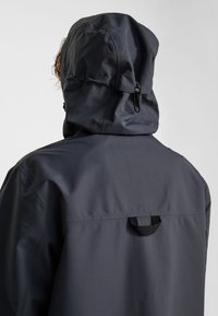 Persoon die een donkergrijze waterdichte jas met verstelbare capuchon draagt, gezien van achteren tegen een effen achtergrond.