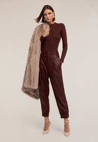 Top a maniche lunghe a coste marrone, scialle in pelliccia sintetica, pantaloni finti in pelle plissé bordeaux e scarpe con tacco alte beige chiare su uno sfondo neutro.