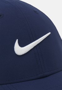 Mørkeblå kasket lavet af tekstureret stof med et fremtrædende hvidt broderet Nike-logo på fronten. Syninger synlige langs kanterne.