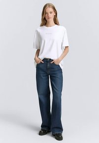 Bela bombažna majica s kratkimi rokavi in subtilnim besedilom, kombinirana s temno modrimi zvončastimi jeans hlačami, ki imajo klasičen dizajn s petimi žepi in zanke za pas.