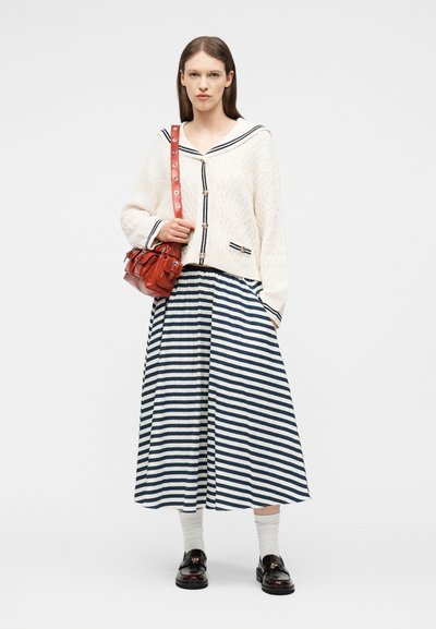 Γυναίκα που φοράει λευκό πλεκτό ζακέτα με μπλε ραφ λεπτομέρειες, φούστα με μπλε και λευκές ρίγες, λευκές κάλτσες, μαύρα μοκασίνια και κρατά κόκκινη δερμάτινη τσάντα.