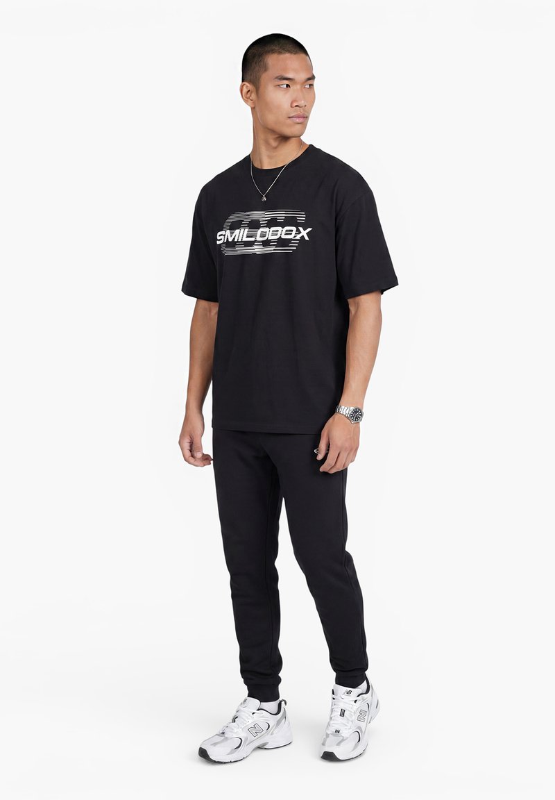 Mann steht, trägt schwarzes Grafik-T-Shirt, schwarze Hose, weiße Sneakers, Uhr und Halskette, blickt nach links vor weißem Hintergrund.