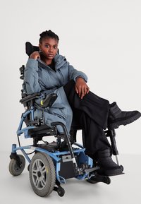 Párnázott szürke kabát kapucnival, fekete bő szárú nadrág és fekete csizmák felett viselve. Kék elektromos kerekesszék nagy kerekekkel és látható vezérléssel.