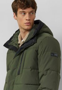 Groene gewatteerde jas met een rits aan de voorkant, zwarte capuchon en zijzakken. Heeft een logo-opdruk op de linkermouw en een gestructureerde stof.