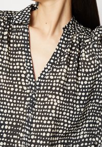 Black blouse with white polka dot pattern, featuring a collar, button-up front, and flowing fabric with pleated details.