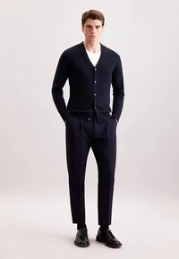 Cardigan blu navy con scollo a V, chiusura con bottoni sul davanti e maniche lunghe, abbinato a pantaloni sartoriali coordinati e scarpe eleganti nere.