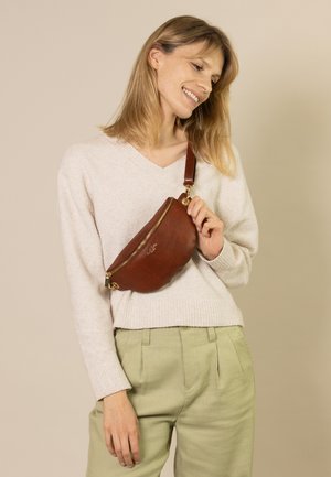 Braune Leder-Gürteltasche mit texturierter Oberfläche, goldfarbenem Reißverschluss und abnehmbarem Trageriemen, getragen über einem cremefarbenen Pullover und hellgrünen Hosen.