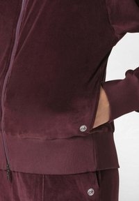 Giacca zip-up in velluto borgogna con polsini e orlo a coste, dotata di tasche laterali e un piccolo logo sullmanica sinistra.